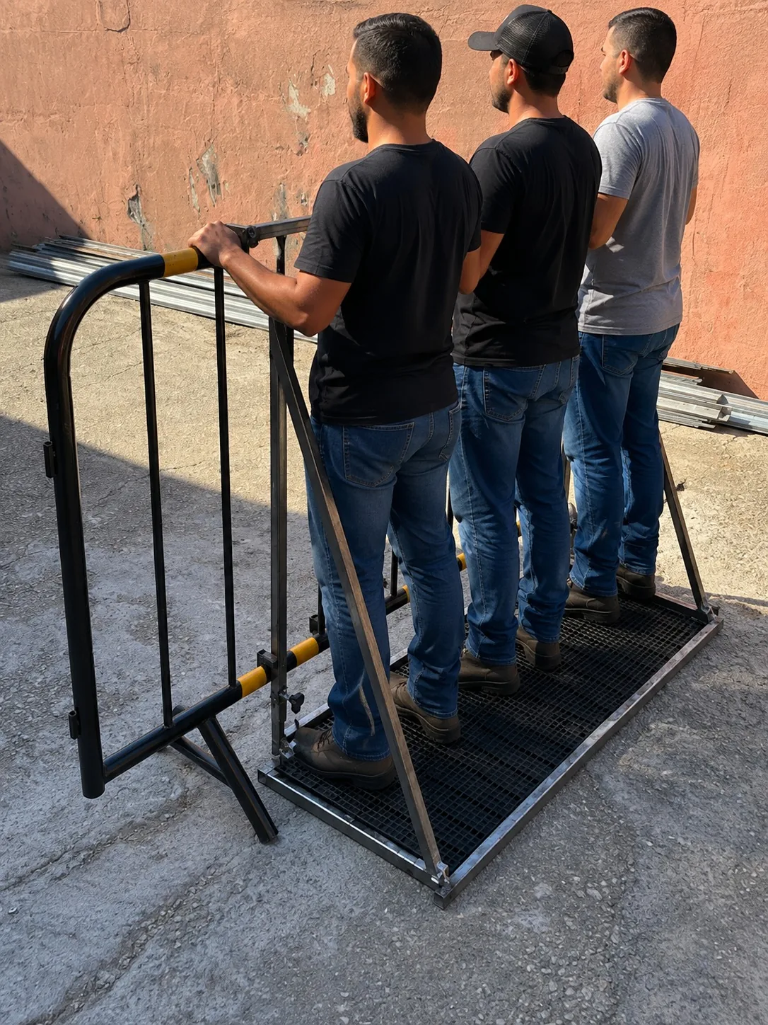 Barricada antitombamento presa a uma grade de isolamento preta com faixas amarelas, com três pessoas em pé sobre o piso